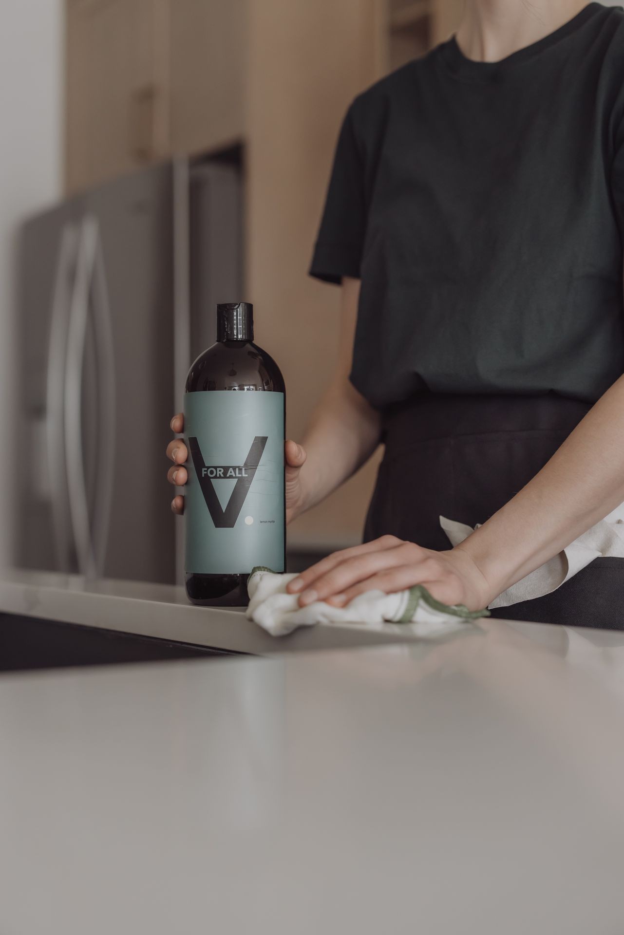 Person wiping a countertop while holding a bottle labeled 'For All' in a kitchen setting.