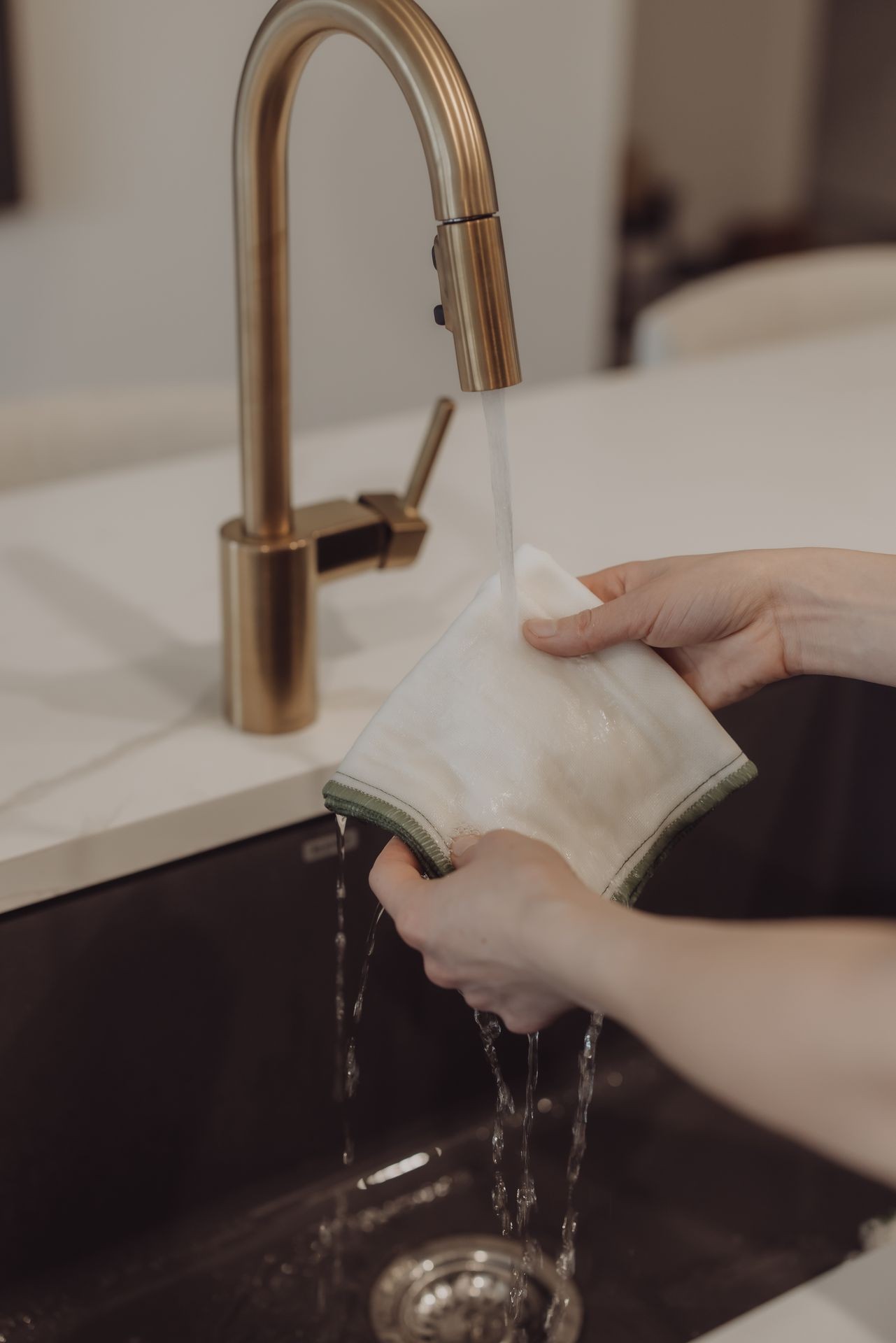 Hands rinsing a white cloth under a bronze kitchen faucet with water flowing over a sink.