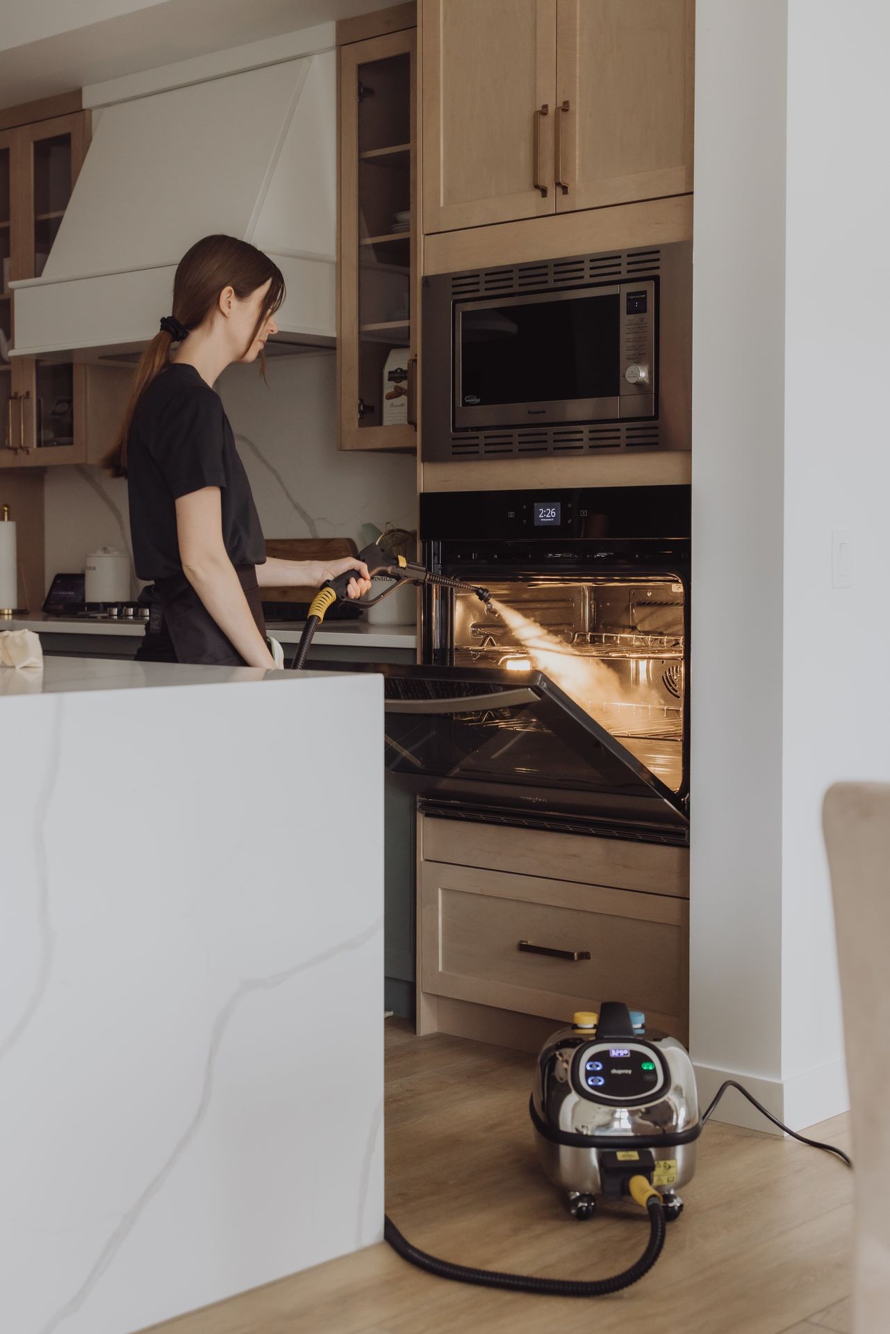 Person cleaning an open oven with a steam cleaner in a modern kitchen setting.