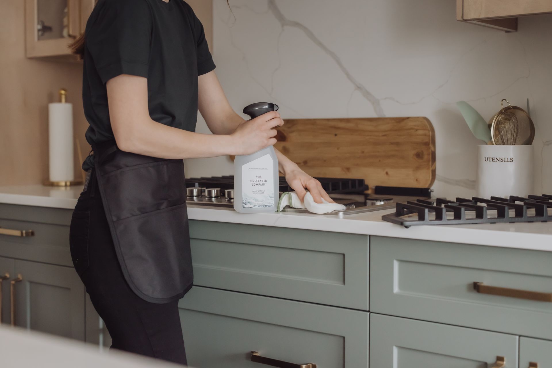 Person cleaning a kitchen countertop with a spray bottle and cloth, next to a gas stove.